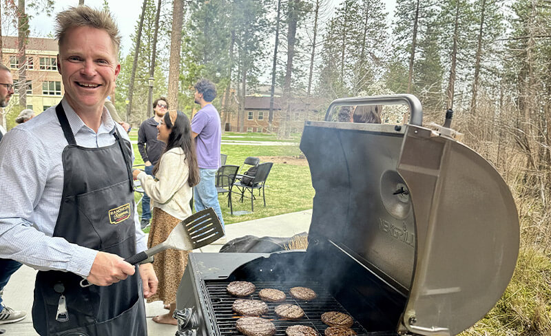 A person grilling burgers outdoors, smiling at the camera, with people and trees in the background.