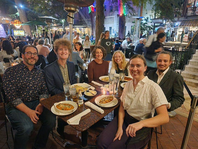 Six students sit around a restaurant table outdoors, sharing a meal and smiling at the camera.