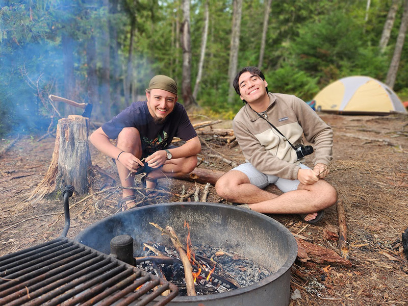 Two students build a campfire in a campground with a tend in the background.