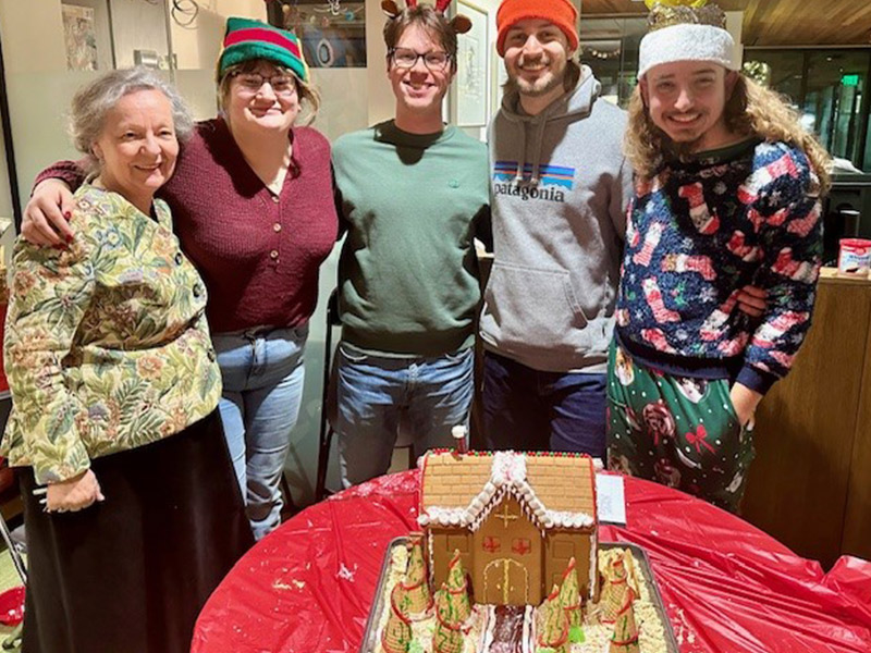Four students in festive hats and sweaters stand with a professor around a table with a gingerbread house on it.