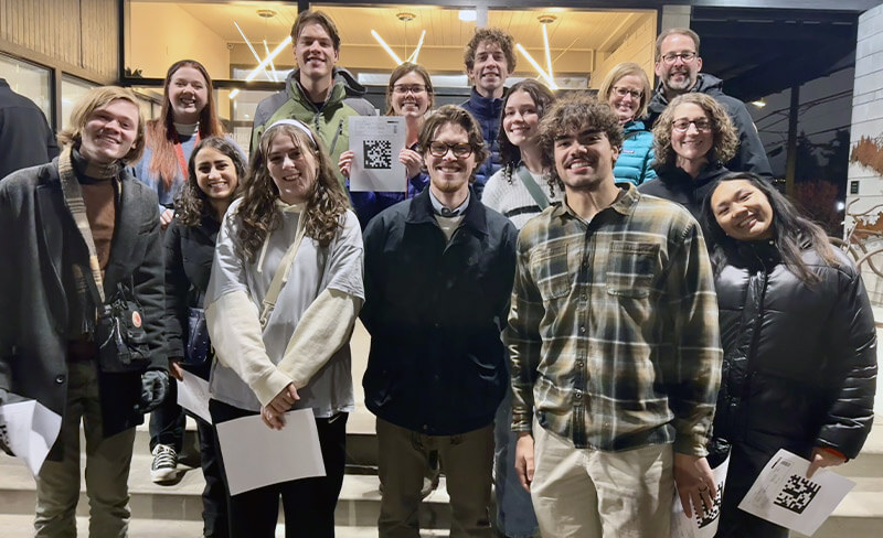 A group of people smiling and posing together outside a building, some holding papers with QR codes.