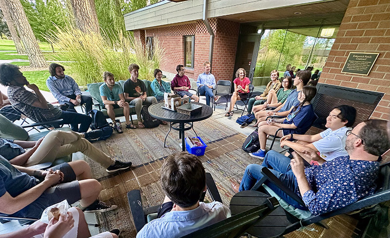 A group of people sits in a circle on a patio, engaged in conversation near a brick building.