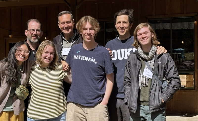 Seven people standing close together outdoors, smiling at the camera on a sunny day