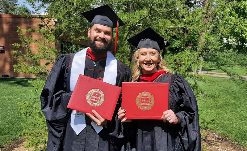 Two graduates in regalia hold up their diplomas and smile at the camera on a sunny day.