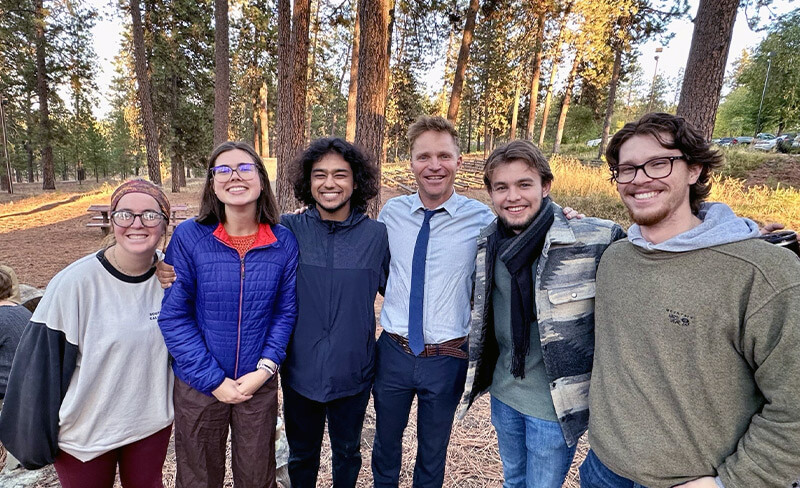 Six people standing close together and smiling outdoors among tall pine trees in a forested area.