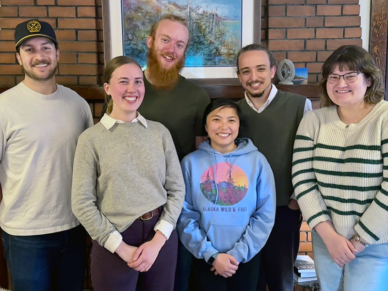 Six students gather in front of a brick interior wall for a photo.