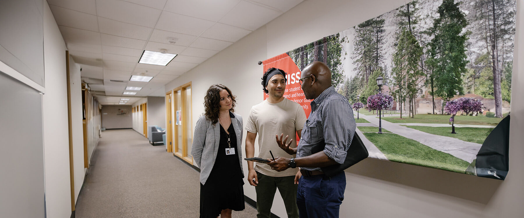 Three people converse in a brightly lit hallway with a campus poster on the wall behind them.