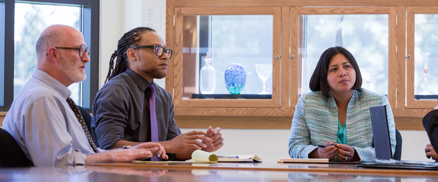 Three professionally dressed adult students sit on one side of a table. They look toward someone speaking out of the frame.