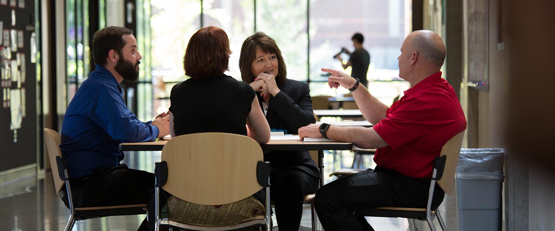 Four adult students sit together at a small, round table. One student talks and points while the others listen.