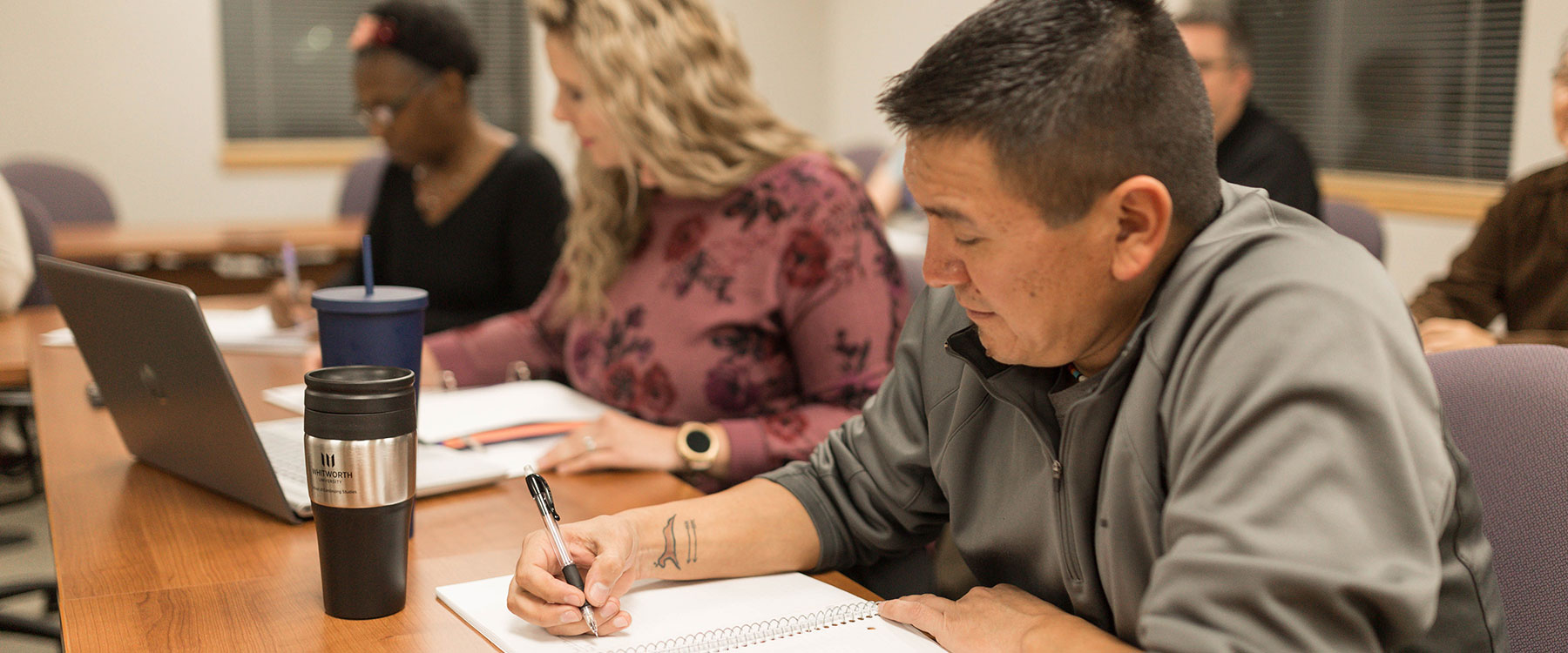 Adult students sitting at a table in a classroom, working on assignments in notebooks and laptops.