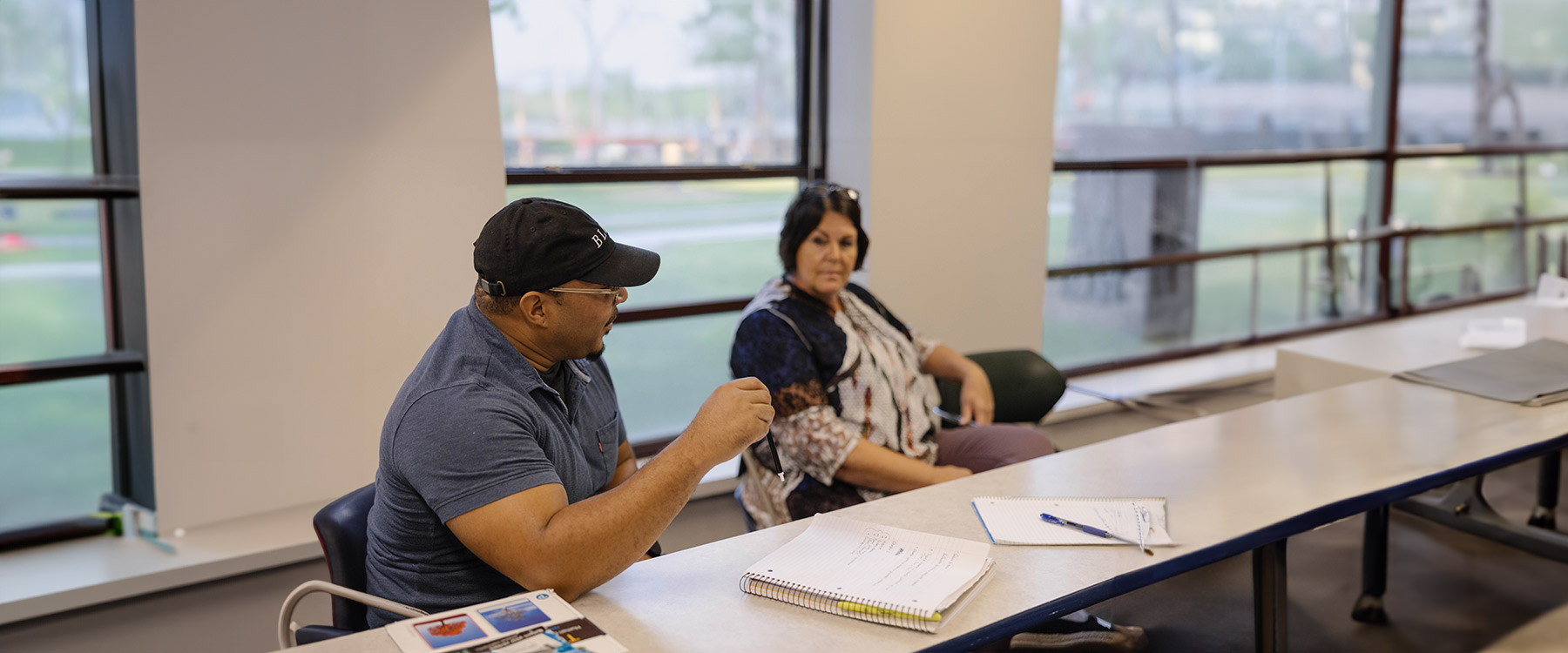 Two adults students sit at a classroom table next to large windows, engaged in discussion.
