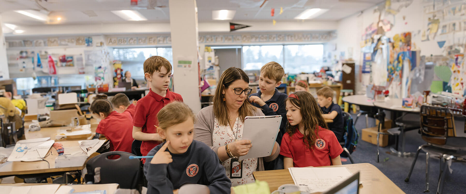 A teacher reads a notebook surrounded by inquisitive elementary students in a private school classroom.