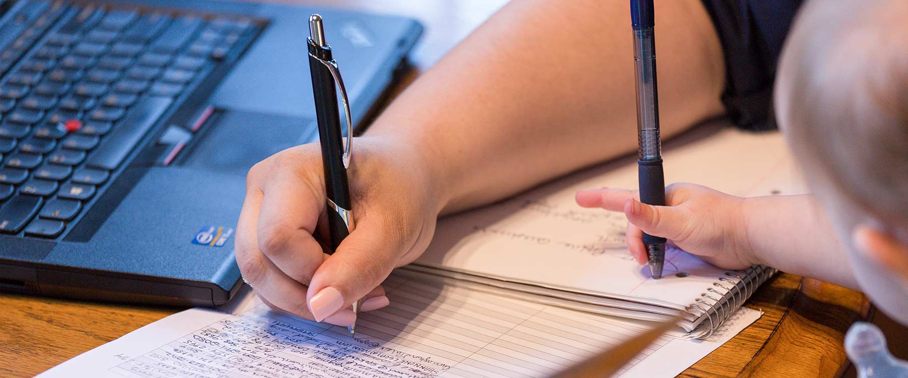 Close-up of two hands, an adults and a toddlers. The parent is writing in a notebook and the child copies.