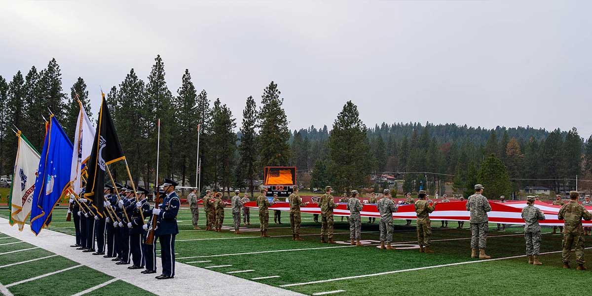 Military personnel hold a large American flag on a football field as a color guard stands at attention.