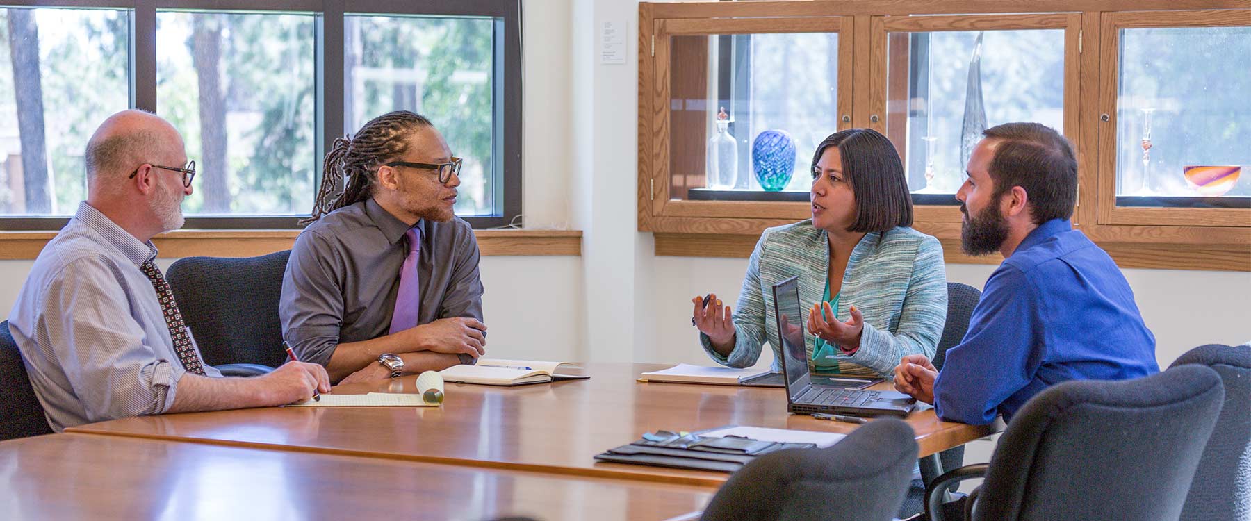 Four professionally dressed adults sit around a long table. One speaks while the others listen.