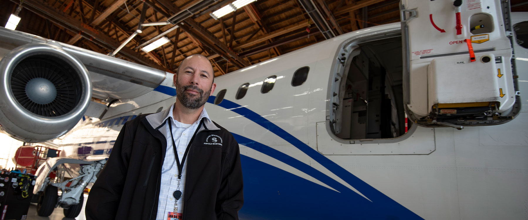 An adult wearing a badge stands in front of a blue and white airplane inside an aircraft hangar.
