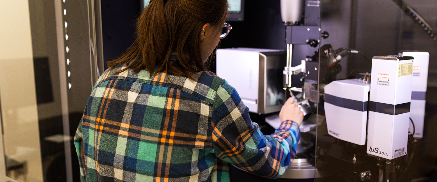 A student in a plaid shirt operates scientific equipment in a laboratory setting.