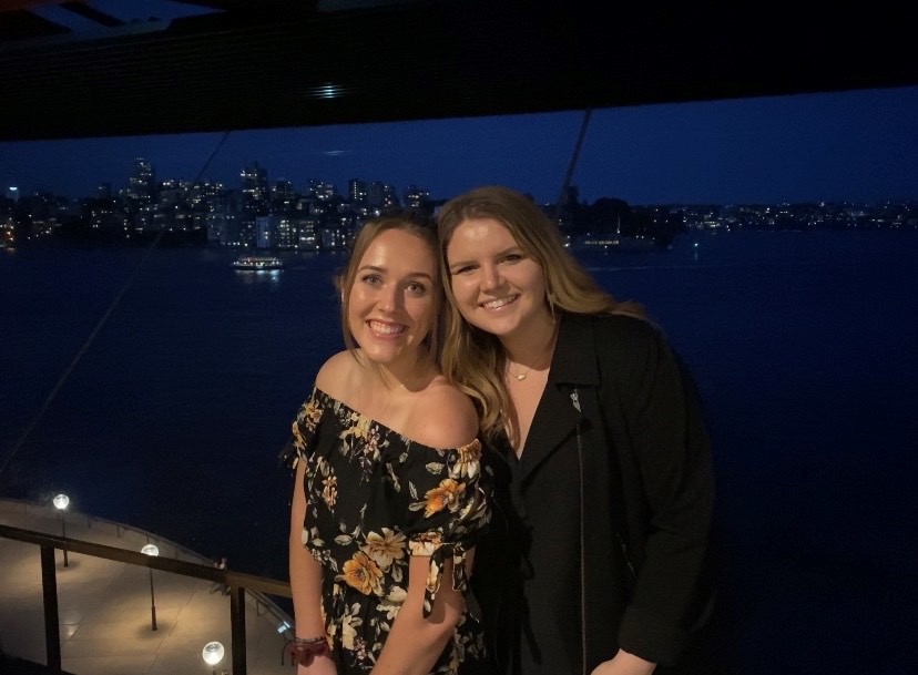 Two young adults pose for a photo at night with a city skyline and body of water behind them.