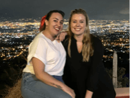 Two young adults post for a photo at dusk with a cityscape behind them.