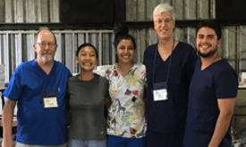 Five people, wearing medical scrubs, standing together and smiling inside a metal building.