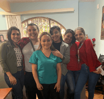 A group of students, one wearing medical scrubs, poses for a photo inside a home.