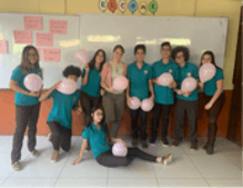 A group of students in matching teal shirts pose with pink balloons in a classroom in front of a whiteboard.