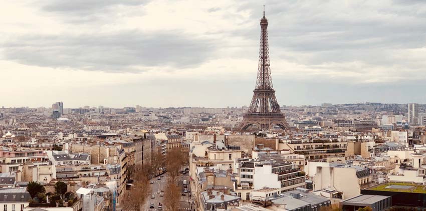 Aerial view of Paris featuring the Eiffel Tower under a cloudy sky. The cityscape is visible with numerous buildings.