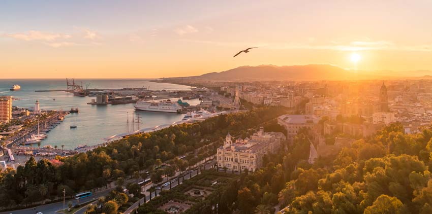 Aerial view of a coastal city at sunset, with a bird flying over, a harbor, ships, and mountains in the background.