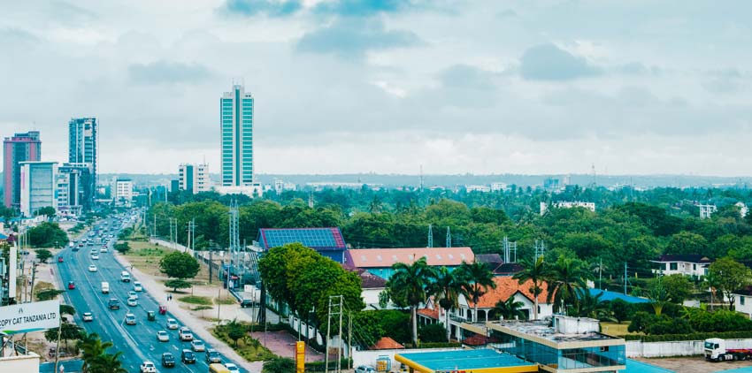 A cityscape featuring a busy road with tall buildings on the left, greenery, and houses on the right under a cloudy sky.