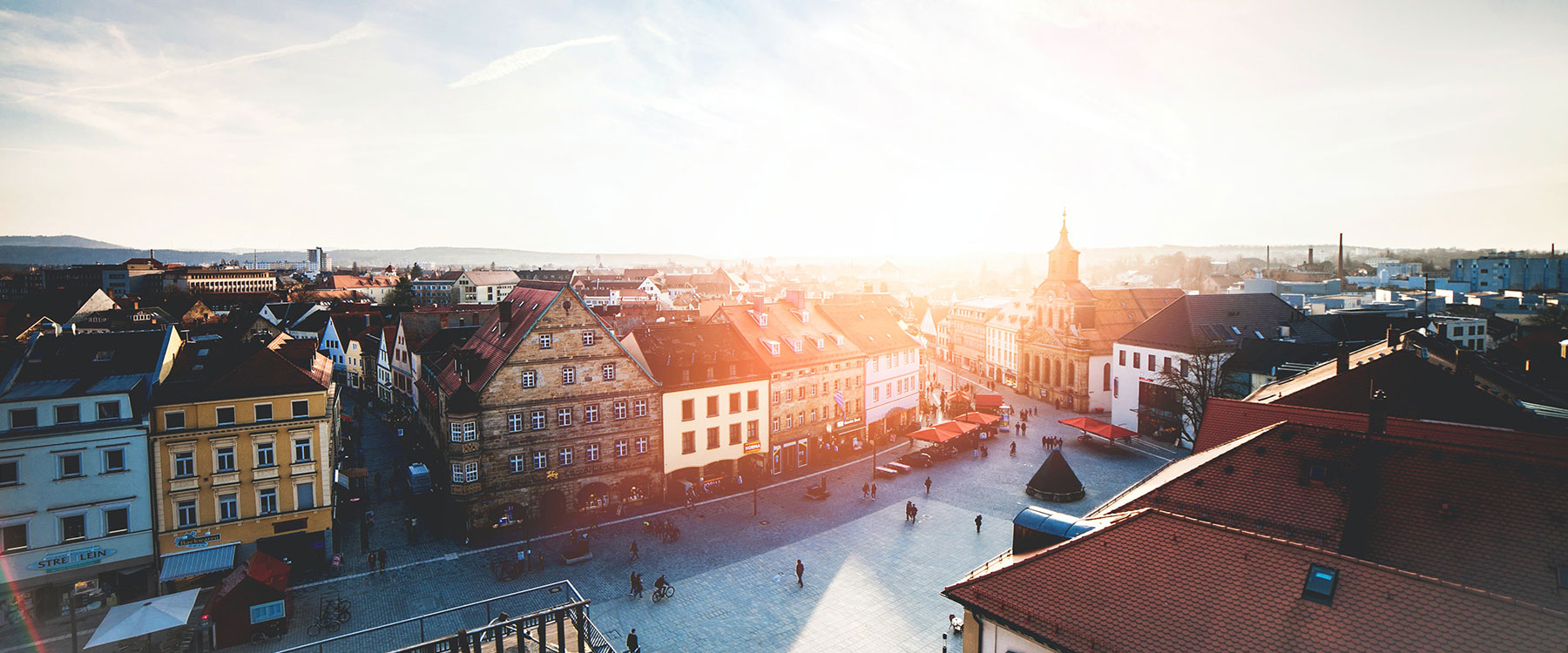 Aerial view of a sunlit European town square surrounded by historic buildings, with light clouds and distant hills.