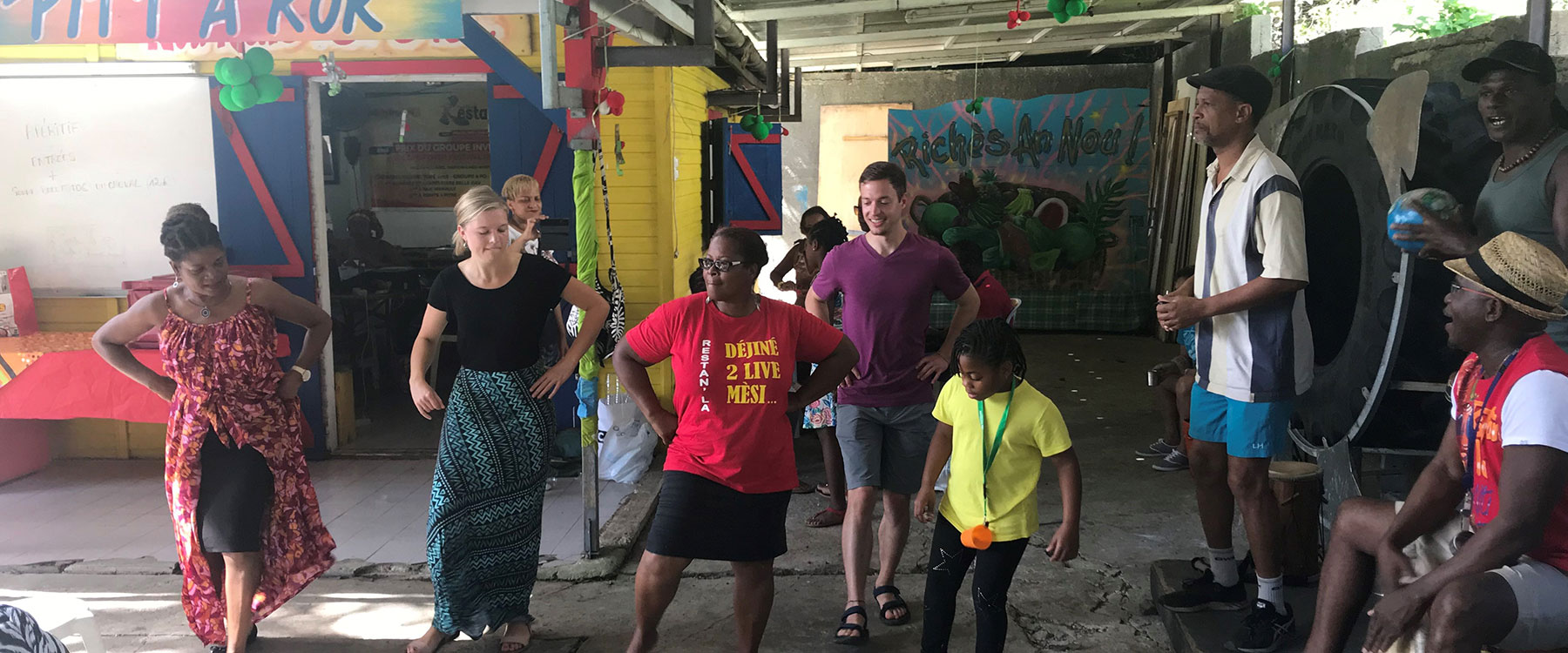 A diverse group of students and locals learn steps to a dance in a colorful, festive environment.
