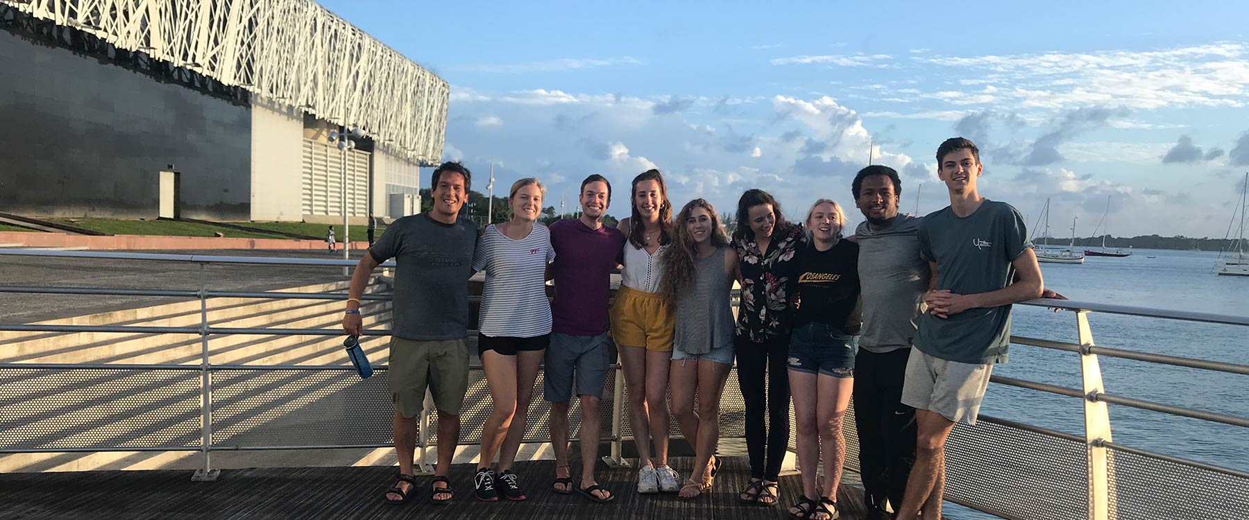 A group of students standing close together, smiling, by a waterfront with buildings and boats in the background.