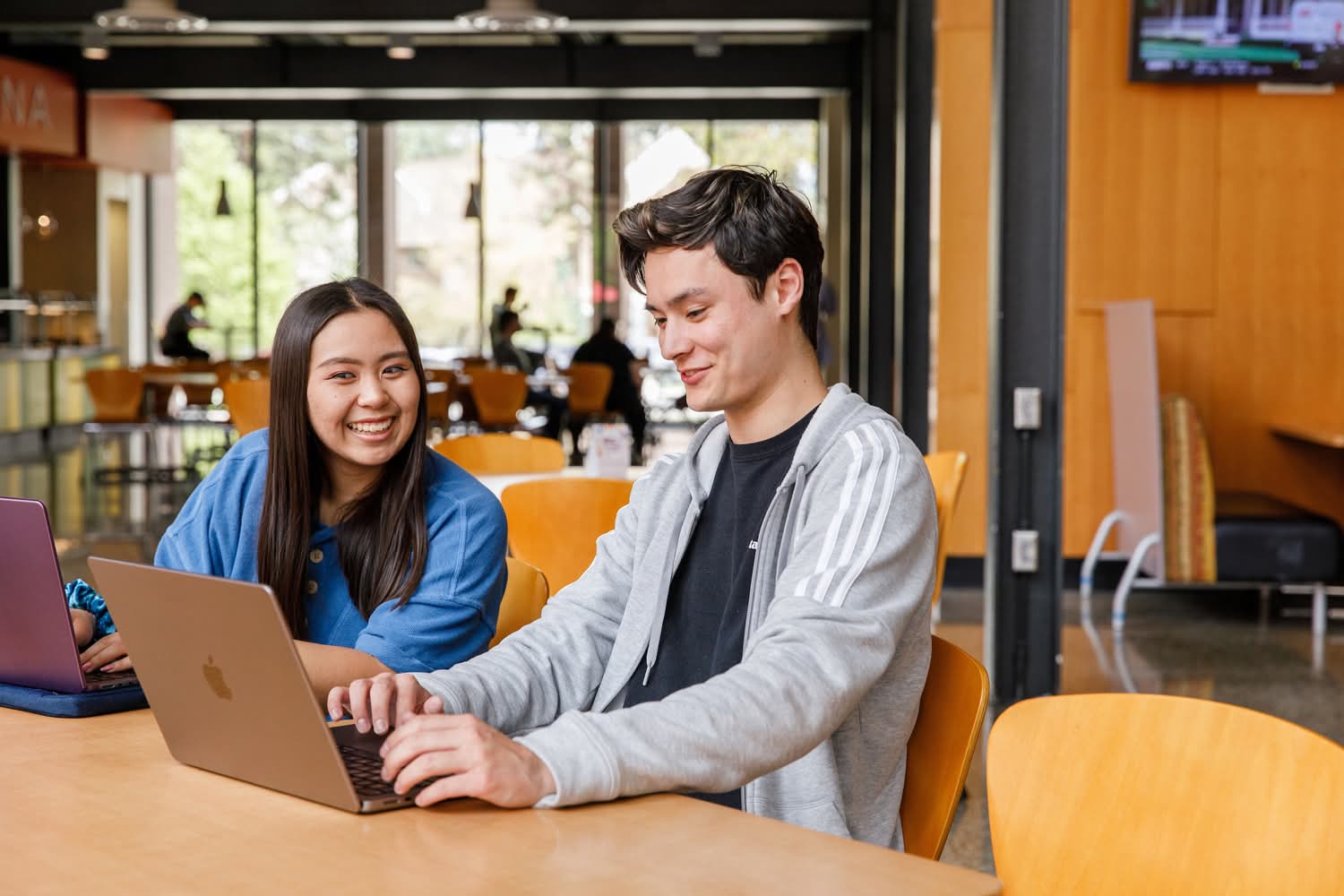 Two students converse at a table, looking at one of their laptop screens.