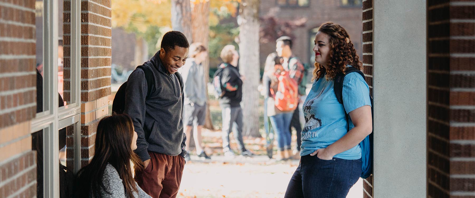 Multiple students, in two small groups, linger outside an academic building, talking.