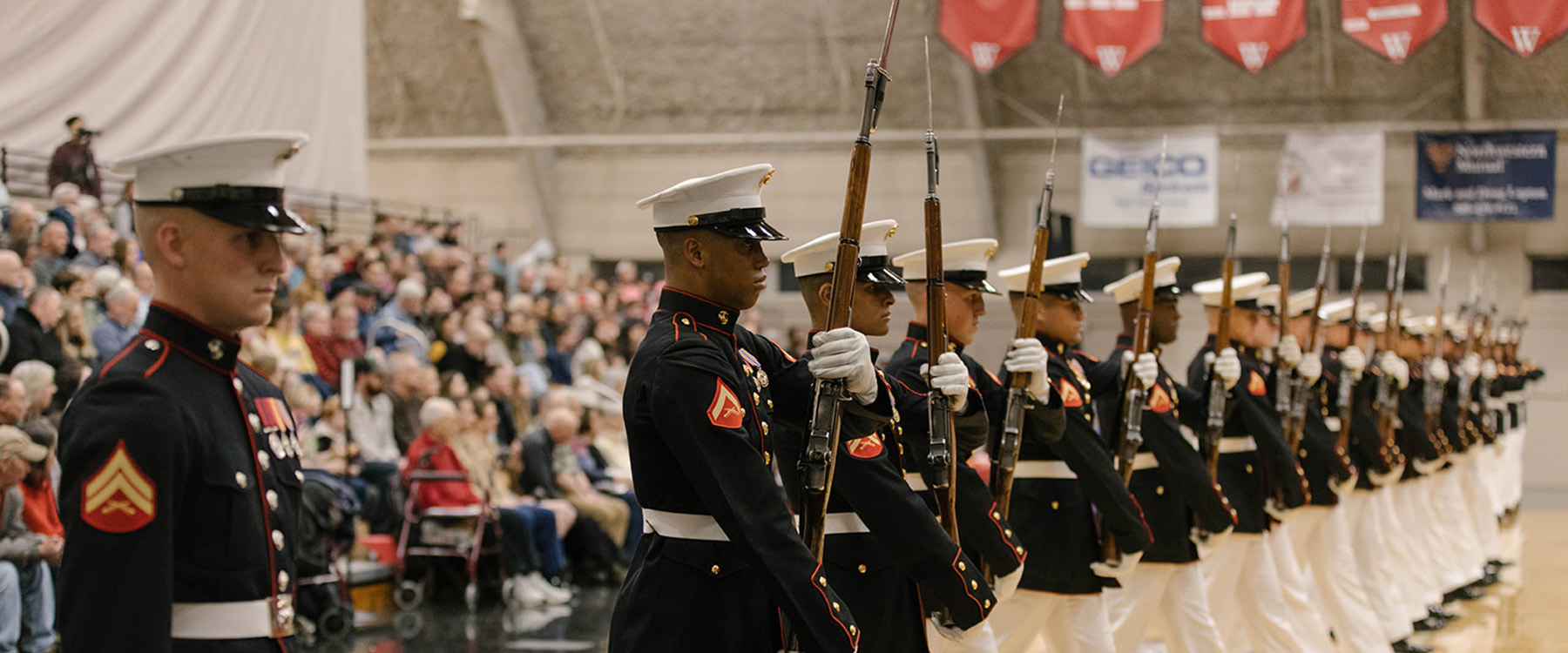 A line of uniformed marines marches in a formal ceremony, holding rifles, while an audience looks on.