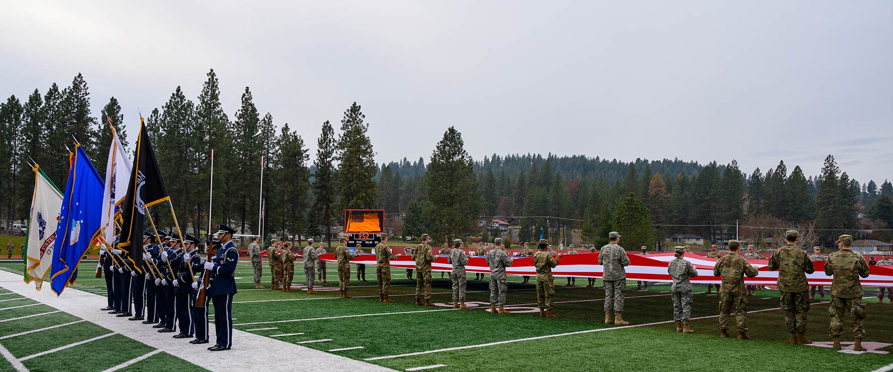 Officers stand with flags on the side of the Whitworth football field, as military members hold a large American flag across the field. 