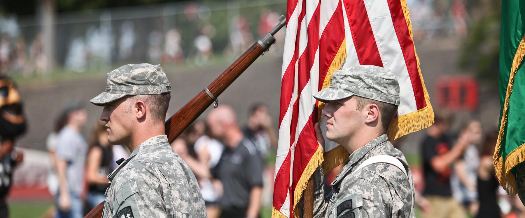 Two ROTC officers stand in a line. One caries a rifle, the other caries the American flag.