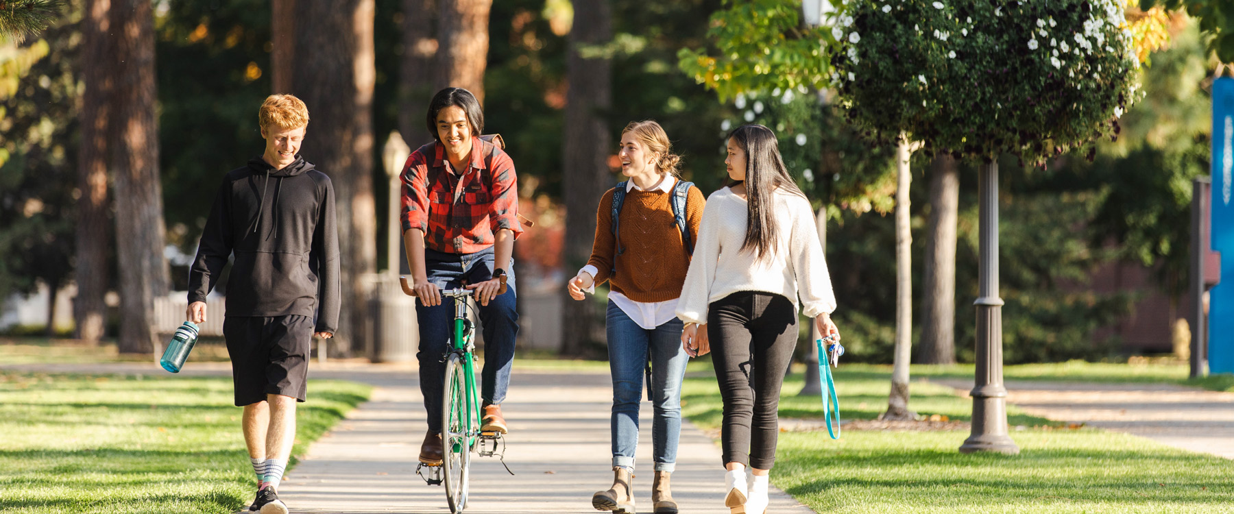 Four students, one on a bike, progress down a campus pathway, smiling and talking.