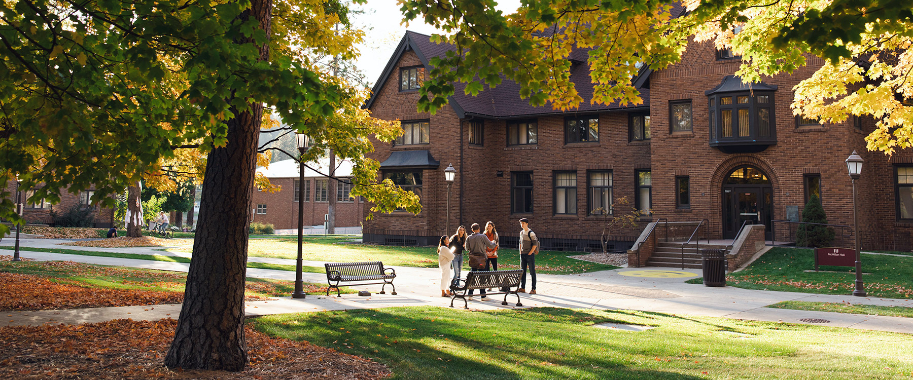 A group of students stands and converses in front of a large brick building on a sunny autumn day.