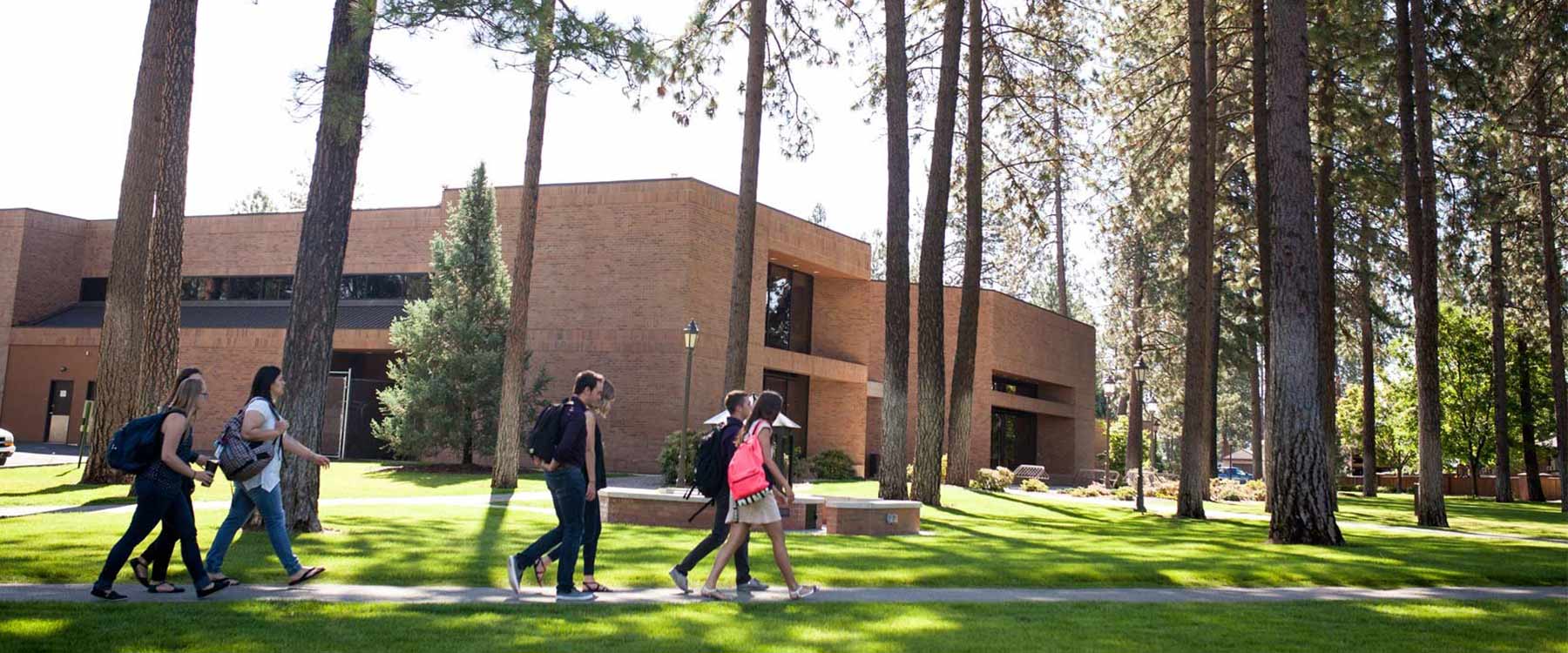 Multiple students wearing backpacks walk past a modern brick academic building beneath pine trees.