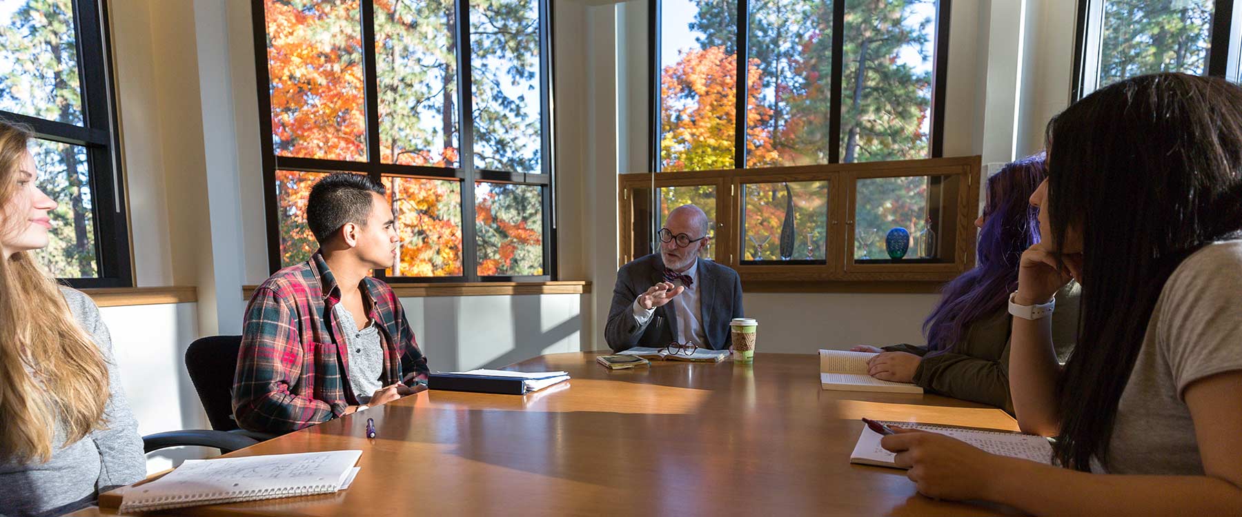 A group of students sit around a table in a window-lined room. A professor speaks at the end of the table.