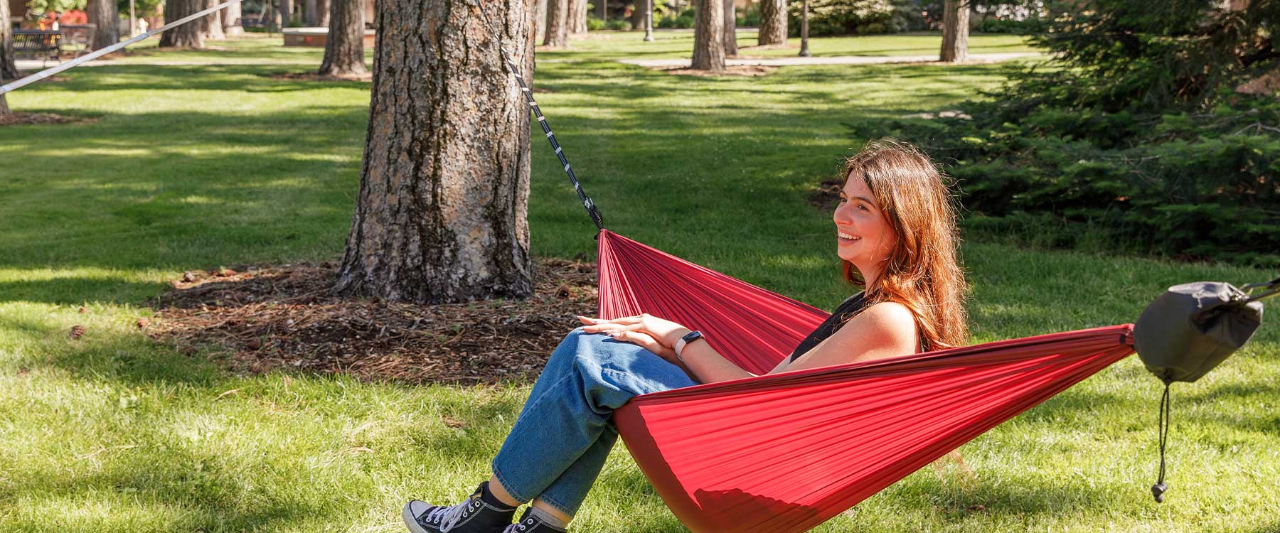 Tall pine trees shade the campus grounds. Between the trees, students sit in hammocks reading.
