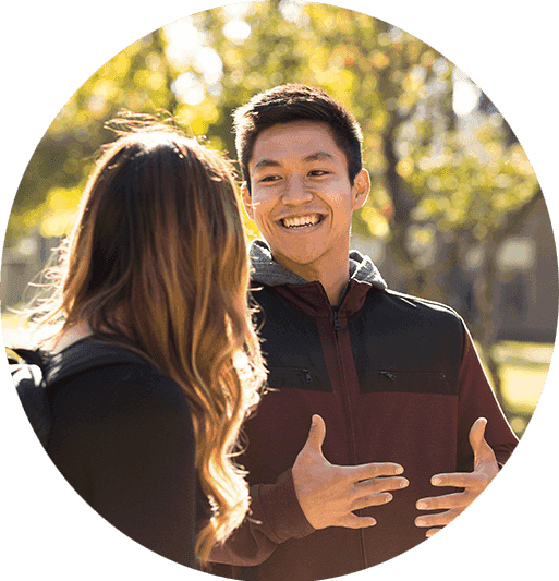 Close up of two students talking, one is seen from behind, the other smiles and gestures enthusiastically.