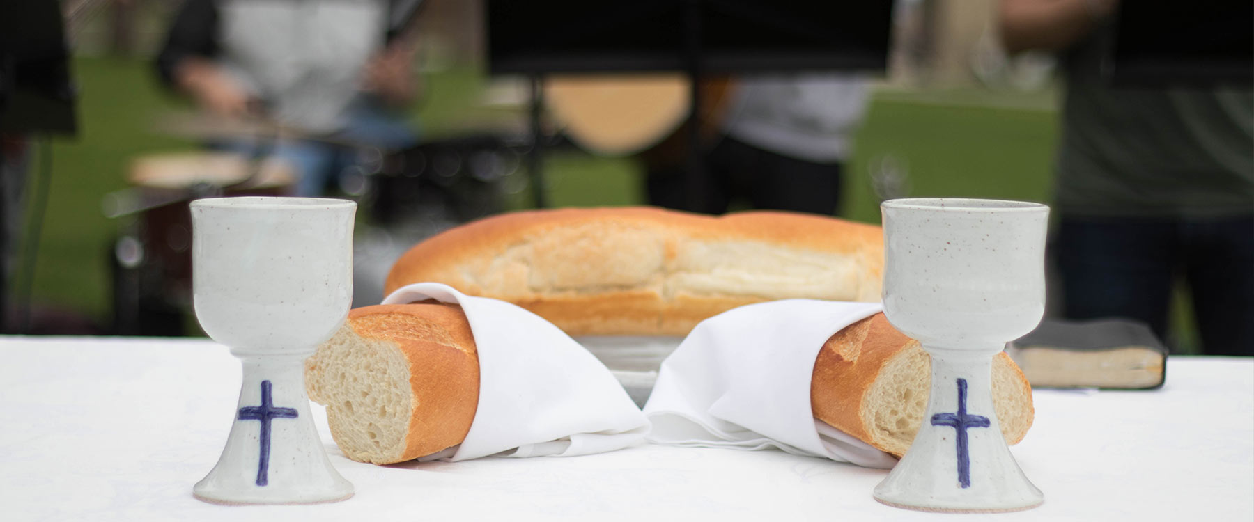 Two ceramic cups, marked with a cross, and a broken loaf of bread sit on a white table cloth.