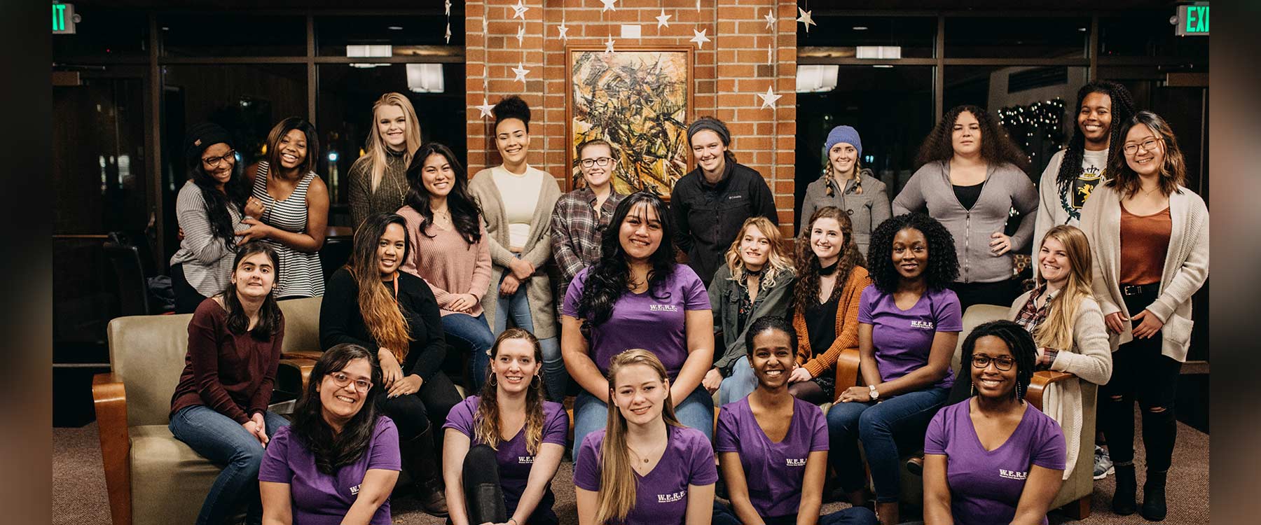 A diverse group of students, some seated, some standing, smile and pose together in a warmly lit room near a brick wall.