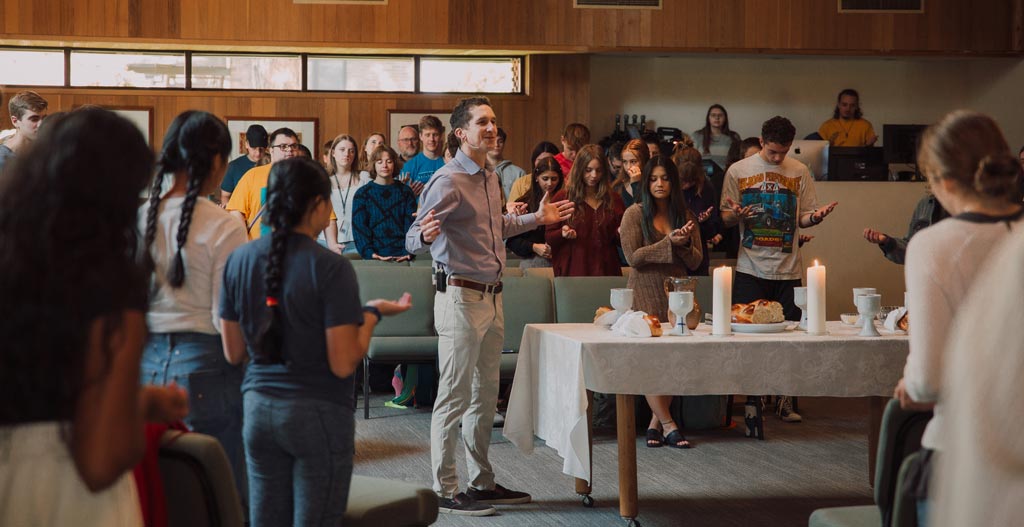 Forrest Buckner lifts his hands and prayers with students over a table set with communion cups and bread.
