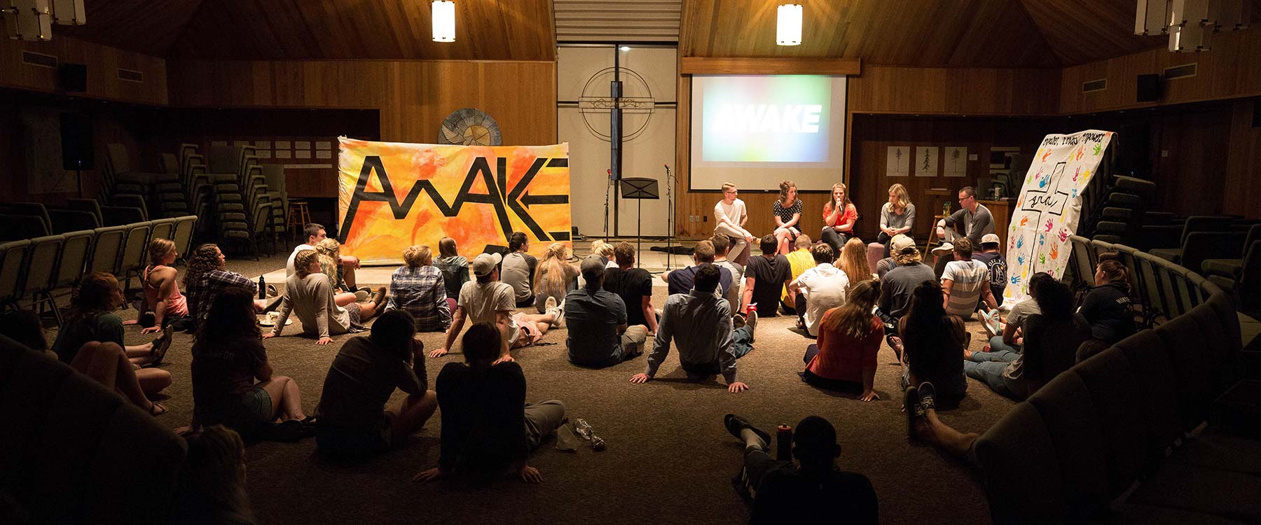A group of students sit on a floor facing a panel of speakers in a dimly lit room with an 