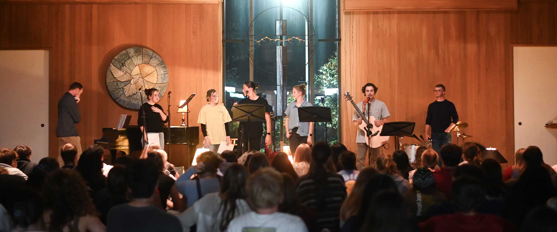 The student worship band, Hosanna, leads a room full of students in worship beneath blue and purple lights.