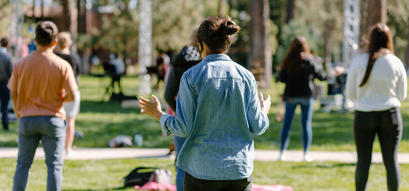 A large group of students stand outdoors on campus, some with arms raised, engaged in a worship service.
