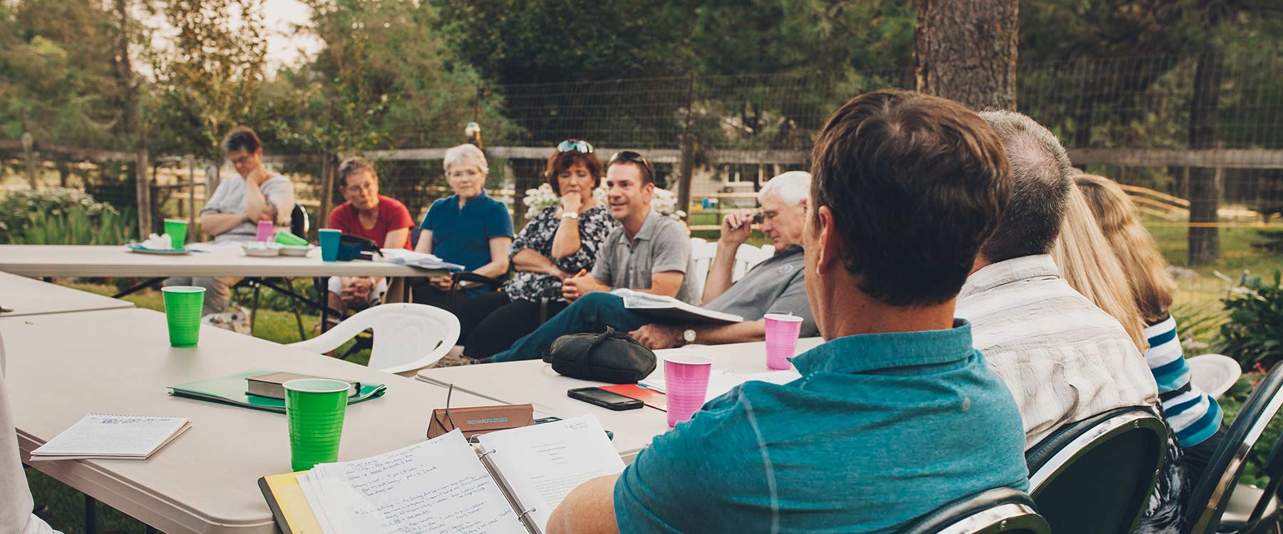 A group of adult students sit outside at tables arranged in a square and listen as one student speaks.